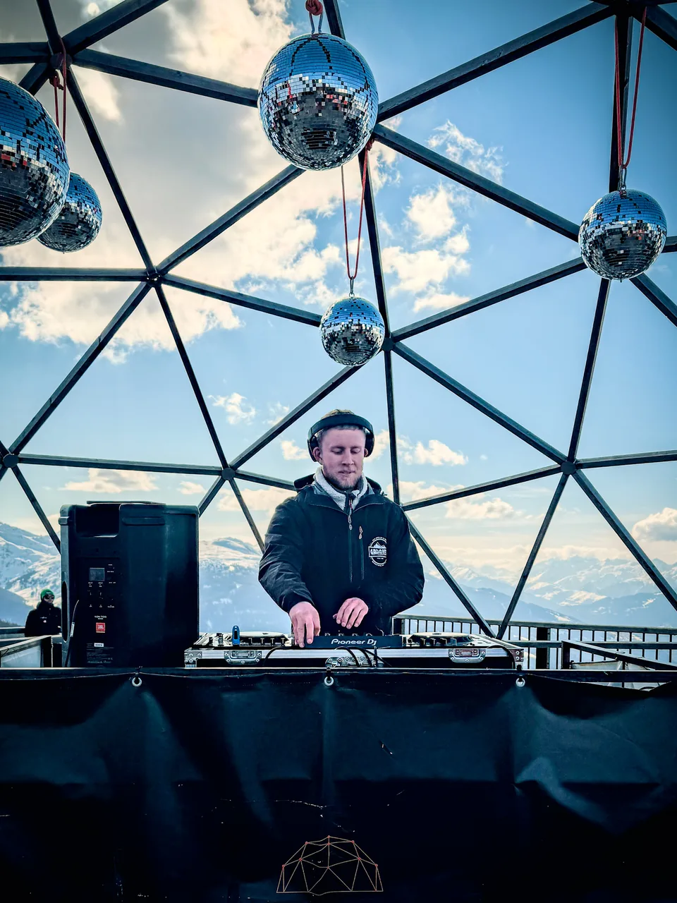 NATUS mixing at a DJ setup beneath disco balls with a snowy mountain backdrop